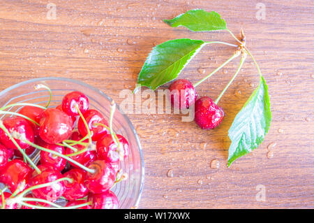 Vista superiore del rosso ciliegie mature in una ciotola di vetro mantenuta su un tavolo di legno in cucina. Alcune ciliegie sul tavolo di legno. Foto Stock