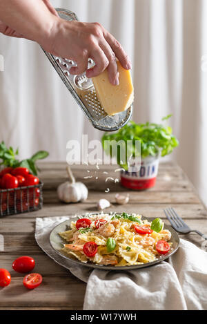 Mani strofinando il formaggio sopra la pasta italiana la piastra. Farfalle in una cremosa salsa di aglio con gamberetti, pomodori ciliegia e basilico su una piastra. Foto Stock