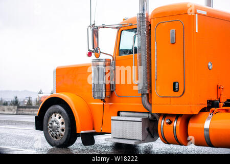 Arancio brillante classica big rig semi carrello con scarico verticale i tubi che trasportano semi rimorchio in esecuzione su vasta multiline strada bagnata in tempo di pioggia Foto Stock