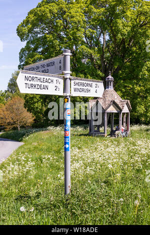 Maytime in Cotswolds - Due femmina i ciclisti nel 1874 pumphouse di fronte all'antica sicomoro sul verde villaggio di Farmington REGNO UNITO Foto Stock