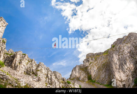 Giovane uomo su un highline sopra sharp scogliere frastagliate. Slackline in montagna con splendidi e verdi pendii e soffici nuvole in background Foto Stock