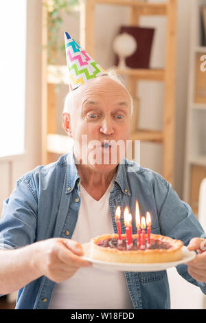 Uomo più anziano soffiando candele sui fatti in casa torta di compleanno mentre si celebra da soli Foto Stock