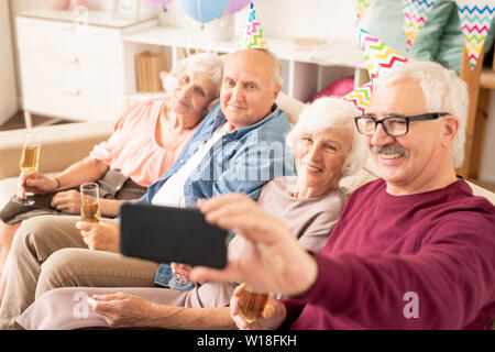 Il gruppo di allegro seniors rendendo selfie sul lettino mentre si sono riuniti per la festa di compleanno a casa Foto Stock