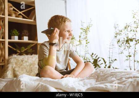 Boy utilizzando diversi gadget a casa. Piccolo modello con smart orologi, smartphone o tablet e cuffie. Rendendo selfie, chating, giochi, guardi i video. Interazione tra bambini e tecnologie moderne. Foto Stock