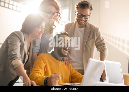 Il gruppo di allegro eccitato giovane interracial colleghi ridere durante la lettura di notizie Internet sul computer portatile Foto Stock