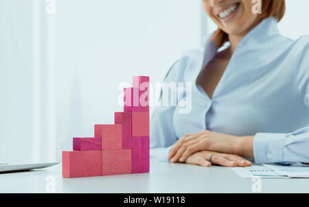 Sorridente imprenditrice costruire con successo un grafico finanziario utilizzando pezzi di legno, essa è fissando il suo lavoro completato Foto Stock
