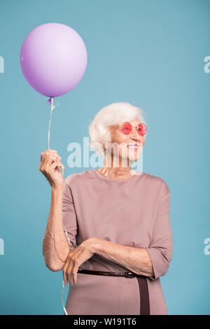 Felice femmina senior in abito elegante azienda palloncino mentre in piedi su sfondo blu Foto Stock