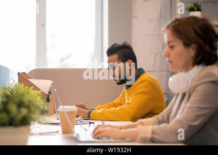 Focus su gravi occupato giovane maschio arabo dipendente in bicchieri seduto alla scrivania e lavorare sul progetto pubblicitario Foto Stock