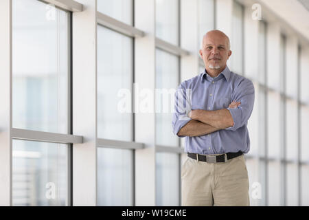 Fiducioso senior imprenditore con bracci ripiegati e un tranquillo sorriso amichevole in piedi nella parte anteriore del vetro sfuggente di windows in un ambiente cittadino Foto Stock