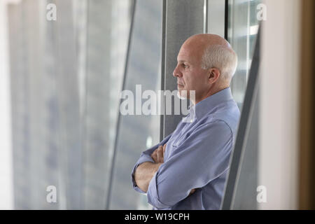 Senior grey-haired man standing nel profondo del pensiero di fronte a una finestra con bracci ripiegati e un deadpan espressione seria, vicino fino in profilo in ambienti interni Foto Stock