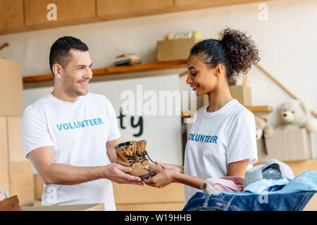 Due giovane e sorridente volontari multiculturale tenendo le scarpe per bambini nel centro di carità Foto Stock