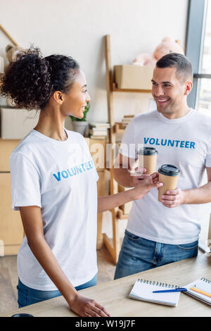 Allegro volontari multiculturale sorridente e parlare tenendo bicchieri di carta Foto Stock