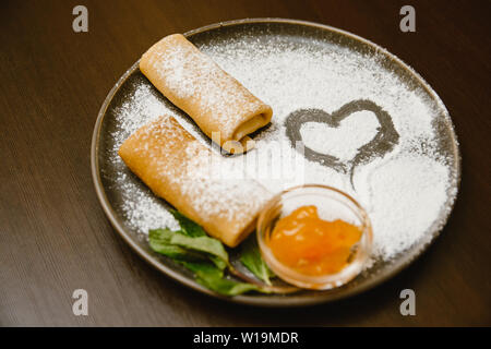 Frittelle rotola su una piastra scuro con zucchero a velo e marmellata e menta. Deliziosi dessert e un cuore di zucchero in polvere. Foto Stock