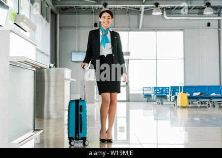 Sorridente African American hostess in piedi con la valigia in aeroporto Foto Stock