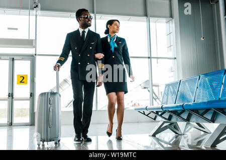 African American pilota hostess e camminare insieme con la valigia in aeroporto Foto Stock