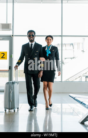 African American pilota hostess e camminare insieme con i bagagli in aeroporto Foto Stock