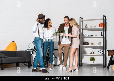 Allegro elegante amici multiculturale tintinnio di bottiglie di birra in casa Foto Stock