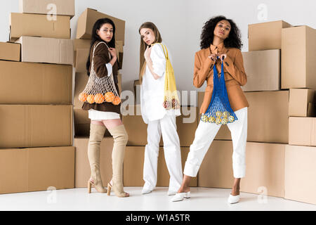 Attraente americano africano donne in piedi vicino a ragazza bionda e trattenere i sacchetti di stringa con frutti su bianco Foto Stock