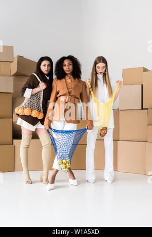 African American girls holding sacchi stringa con frutti nei pressi di donna bionda e scatole su bianco Foto Stock