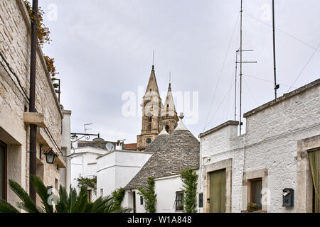 Grande trulli case con tetti conici sulla vecchia strada con piante verdi sulla chiesa medievale di sfondo in Alberobello in Italia. Ci sono v Foto Stock
