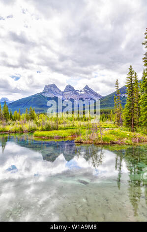 Canmore i famosi tre sorelle picchi di montagna e le loro riflessioni sul Fiume Bow nel sud di Banff gamma delle Canadian Rockies su un nuvoloso S Foto Stock