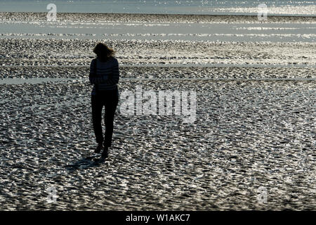 Lonely donna vede nella retroilluminazione come lei cammina sulla sabbia bagnata con la bassa marea, Le Crotoy, Baia di Somme, Somme, Hauts de France, Francia Foto Stock