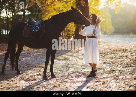 Donna sorridente in abito bianco con bouquet di fiori secchi in piedi a cavallo nel parco. Foto Stock