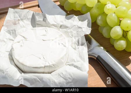 Aprire il pacchetto di carta di tutta la testa di camembert, un coltello e dolce uva verde sotto un sole dalla finestra su un legno marrone dello sfondo. Foto Stock