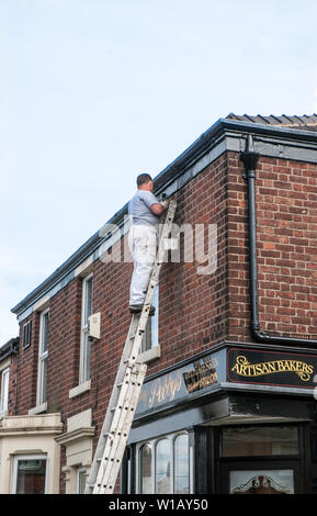 Pittore su scala estensibile pittura nera linea del tetto e grondaie al di sopra di un delicatessen panificatori artigiani shop Foto Stock