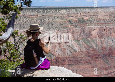 Tourist donna con cappello e zainetto di scattare una foto con un telefono cellulare seduto su di una scogliera in South Rim, il Parco Nazionale del Grand Canyon, Arizona, Stati Uniti d'America Foto Stock