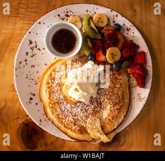 La prima colazione con pancake, sciroppo d'acero, crema, frutta fresca servita su piastra bianca sulla cima di un tavolo di legno Foto Stock