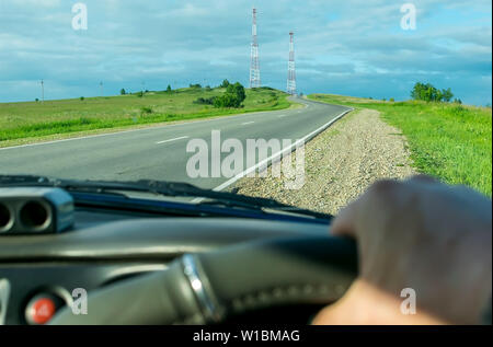 Vista dal lato del conducente della vettura le mani sul volante di sterzo sullo sfondo del paesaggio e salendo su per la collina strada con torri radio Foto Stock