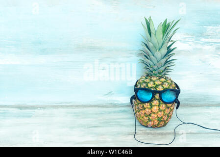 Di ananas maturo con specchio azzurro occhiali da sole e le cuffie nella parte anteriore di un blu rustico in legno sfondo. Foto Stock