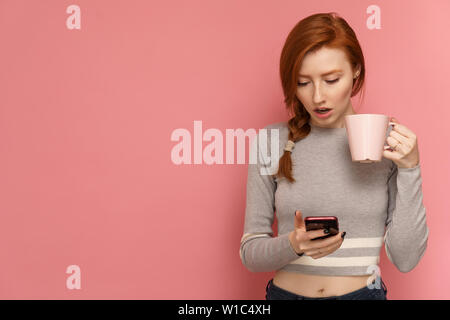 Redhead giovane donna tiene un bicchiere in una mano fissando il suo smartphone in altra parte a bocca aperta Foto Stock