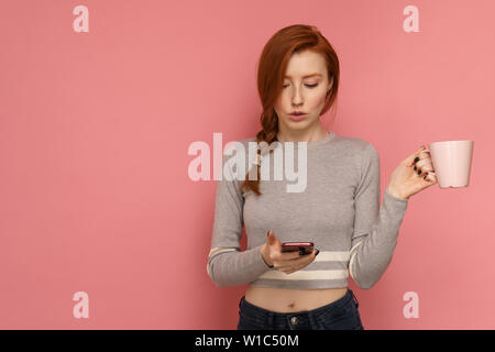 Redhead giovane donna tiene un bicchiere in una mano fissando il suo smartphone in altri invece sorprendentemente Foto Stock