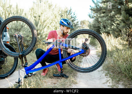 Giovane uomo in sportswear riparazione bicicletta sulla strada forestale durante il periodo estivo Foto Stock
