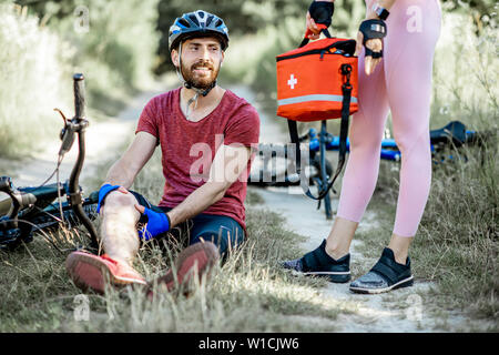 Donna di dare prima un aiuto a un uomo con infortunio al ginocchio durante il ciclismo in mountain bike sulla strada forestale durante il periodo estivo Foto Stock