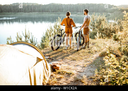 Coppia giovane in piedi con la mountain bike presso il campeggio, viaggiando nella foresta vicino al lago al tramonto Foto Stock
