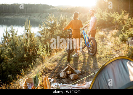 Coppia giovane in piedi con la mountain bike presso il campeggio, viaggiando nella foresta vicino al lago al tramonto Foto Stock