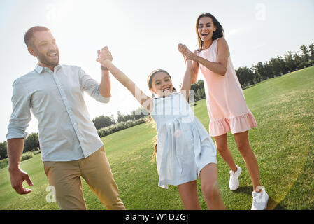 Giochiamo a lunghezza piena di giovani e genitori felici portando la loro figlia e sorridenti mentre spendere tempo insieme all'esterno. Concetto di famiglia Foto Stock