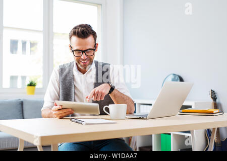 Felice l'uomo che lavora da casa su tavoletta digitale e il computer portatile Foto Stock