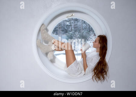 Bella ragazza in bianco l'usura si trova sulla pedana rotonda finestra e caffè drinkig Foto Stock