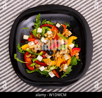 Vista superiore della vitamina insalata di rucola con peperoni e formaggio feta decorate con olive e noci servita nella piastra nera su striped backgro tessili Foto Stock