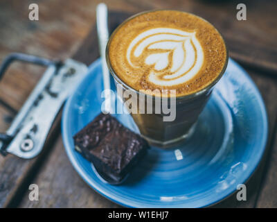 Close-up Piccolo Latte topping caffè con flower art dal latte in vetro di piccole dimensioni con un pezzo di fatti in casa torta brownie su blu piastra ceramica su legno