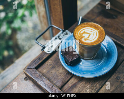 Piccolo Latte topping caffè con flower art dal latte in vetro di piccole dimensioni con un pezzo di fatti in casa torta brownie su blu piastra ceramica sul vassoio in legno su w Foto Stock