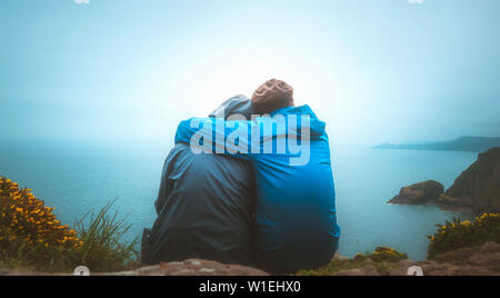 Vista retro o vista posteriore di due giovani amanti seduto sul bordo di una scogliera a guardare il mare su una piovosa e moody giorno. Partnership, all'aperto, tra Foto Stock