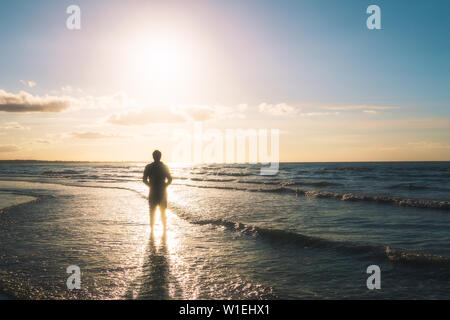 Silhouette di un giovane uomo in piedi sulla riva del mare di fronte al sole. All'aperto, estate, libertà concetto con copia spazio. Foto Stock