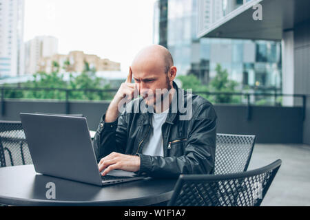 Il pensiero allettante successo per adulti calvo uomo barbuto in giacca nera con il computer portatile in street cafe presso la città Foto Stock