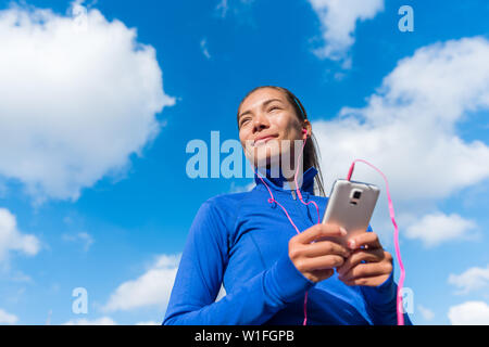 In funzione dello stile di vita della donna ascoltando musica fitness playlist sul telefono intelligente che indossa gli auricolari o cuffie intrauricolari. Andando femmina per eseguire la formazione in sotto il cielo blu. Runner ragazza all'aperto. Foto Stock