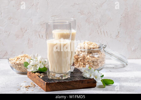 Due bicchieri con Latte di avena e fiori di Apple sulla scheda bruciata e fiocchi di avena su sfondo chiaro. Foto Stock